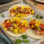Three assembled salmon tacos with pineapple salsa on a baking sheet.