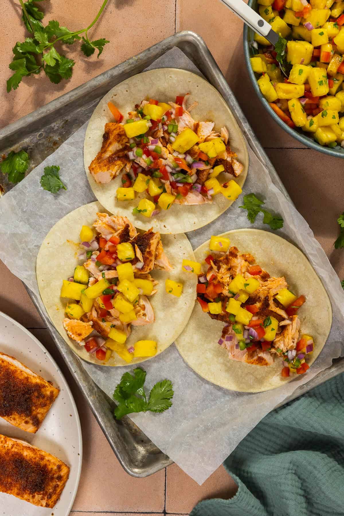 Three corn tortillas on a baking sheet topped with salmon and pineapple salsa.