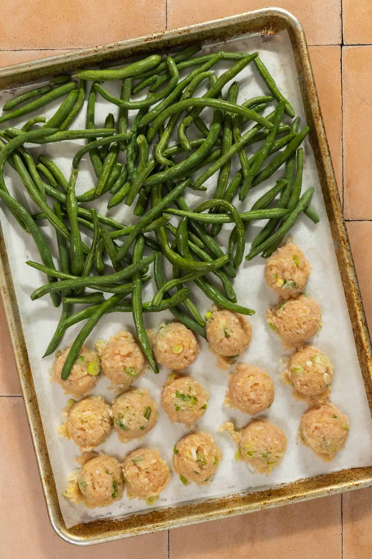 A parchment lined baking sheet with trimmed green beans and chicken meatballs prior to baking.