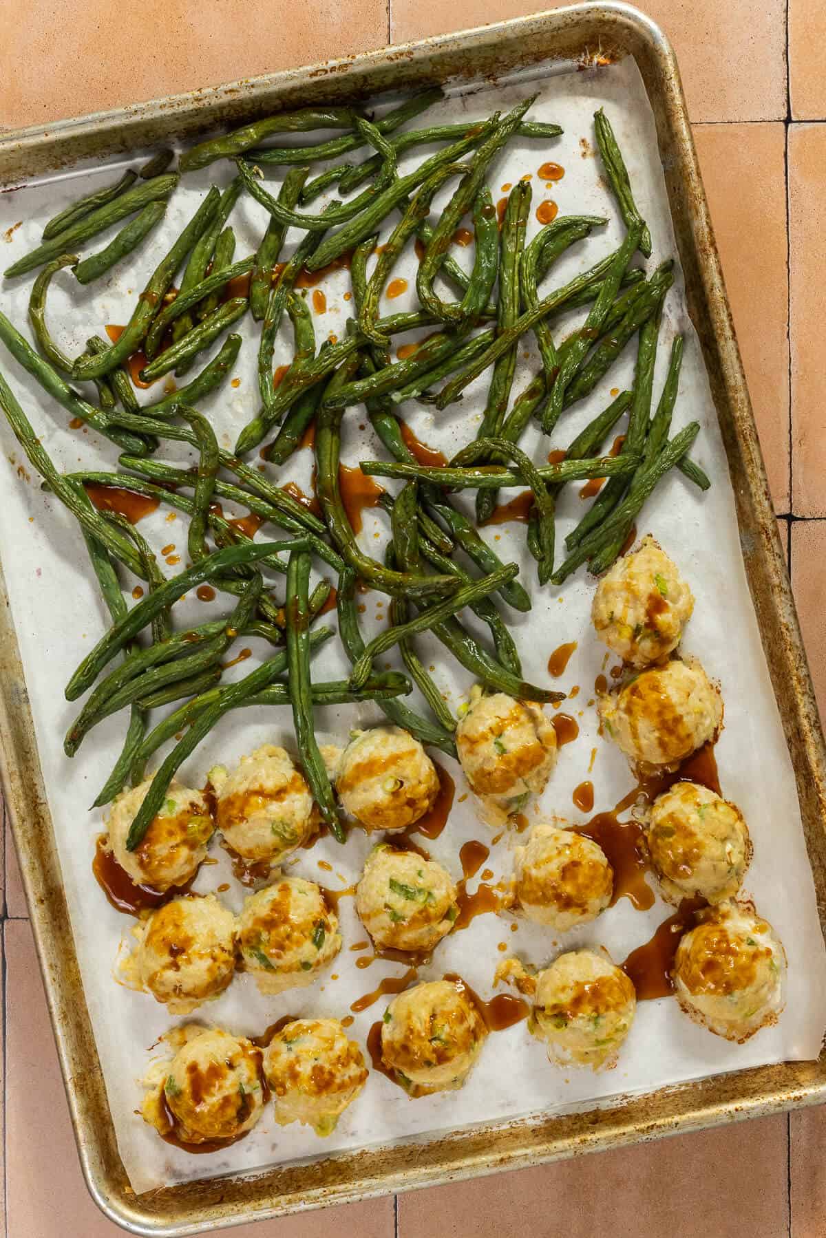 A baking sheet with baked meatballs and green beans, drizzled with sauce.