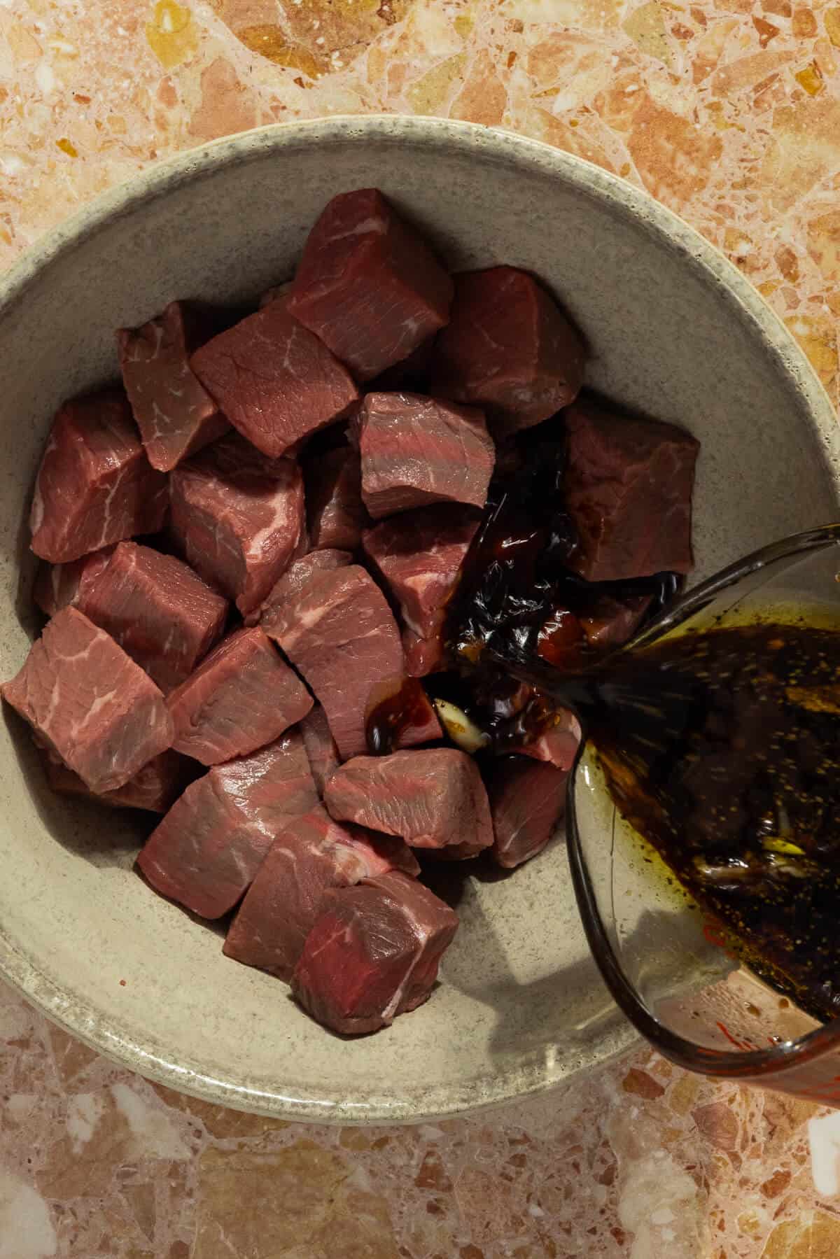 A large mixing bowl with the marinade being poured onto the beef cubes.