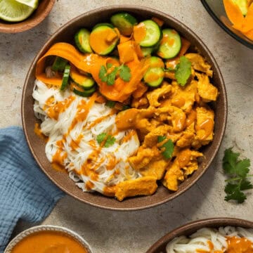 A chicken satay bowl with satay chicken, rice noodles, cucumber carrot salad, and satay sauce drizzled on top.