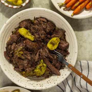 A bowl of Mississippi pot roast served alongside pepperoncini, roasted carrots, and roasted potatoes.