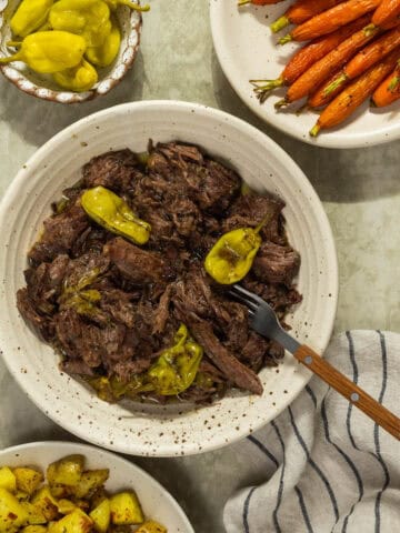 A bowl of Mississippi pot roast served alongside pepperoncini, roasted carrots, and roasted potatoes.