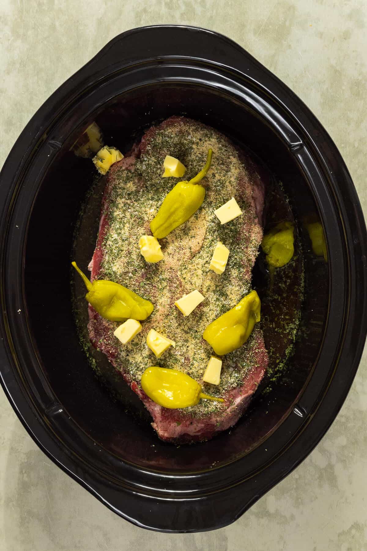 Crock pot insert with pot roast topped with seasonings, cubes of butter, and pepperoncini peppers.
