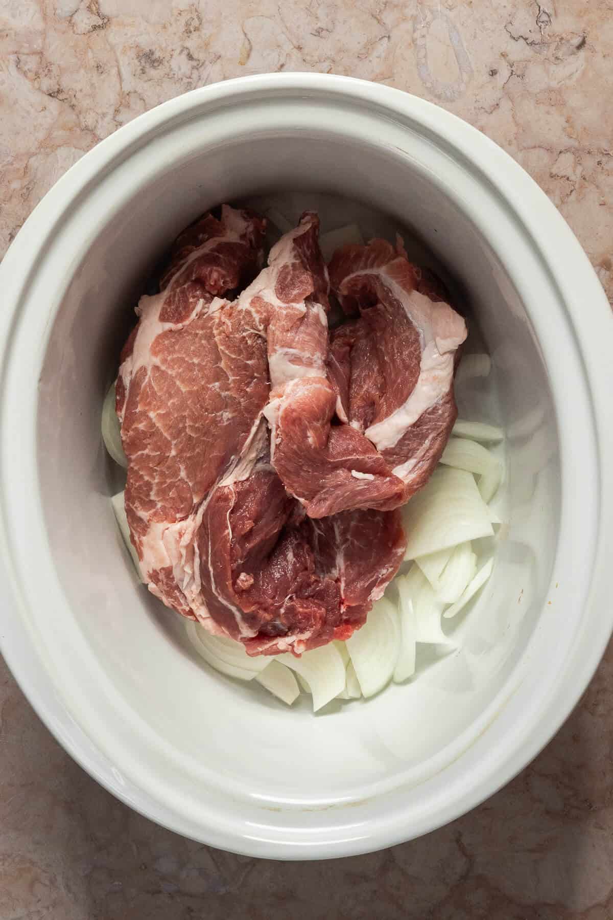 A crockpot insert with sliced onion and a raw pork butt/shoulder prior to seasoning.