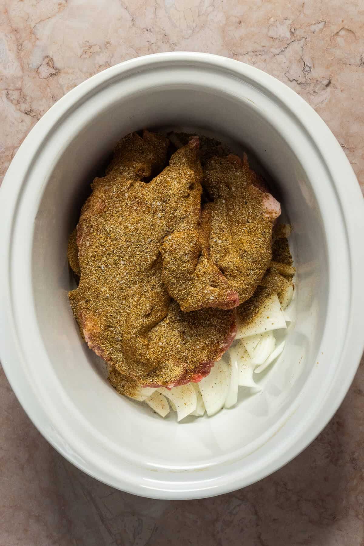 A crockpot insert with sliced onion and a raw pork butt/shoulder after seasoning.