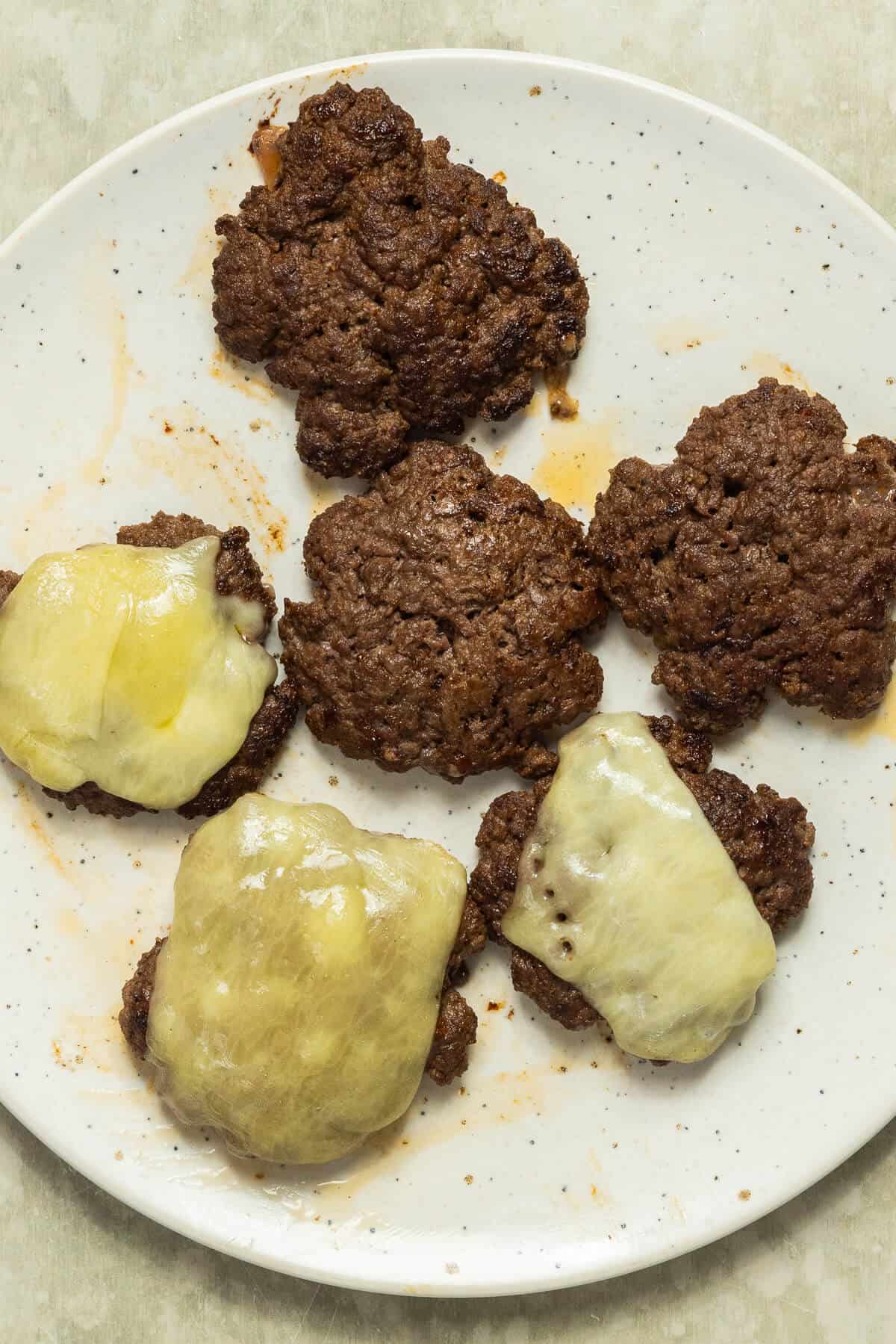 A plate with six cooked smashburgers, some with melted cheese and some without.
