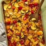 A baking sheet and wooden serving spoon with greek sheet pan chicken and vegetables.
