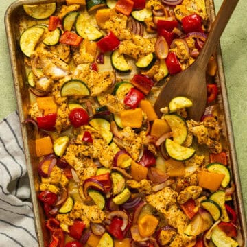 A baking sheet and wooden serving spoon with greek sheet pan chicken and vegetables.