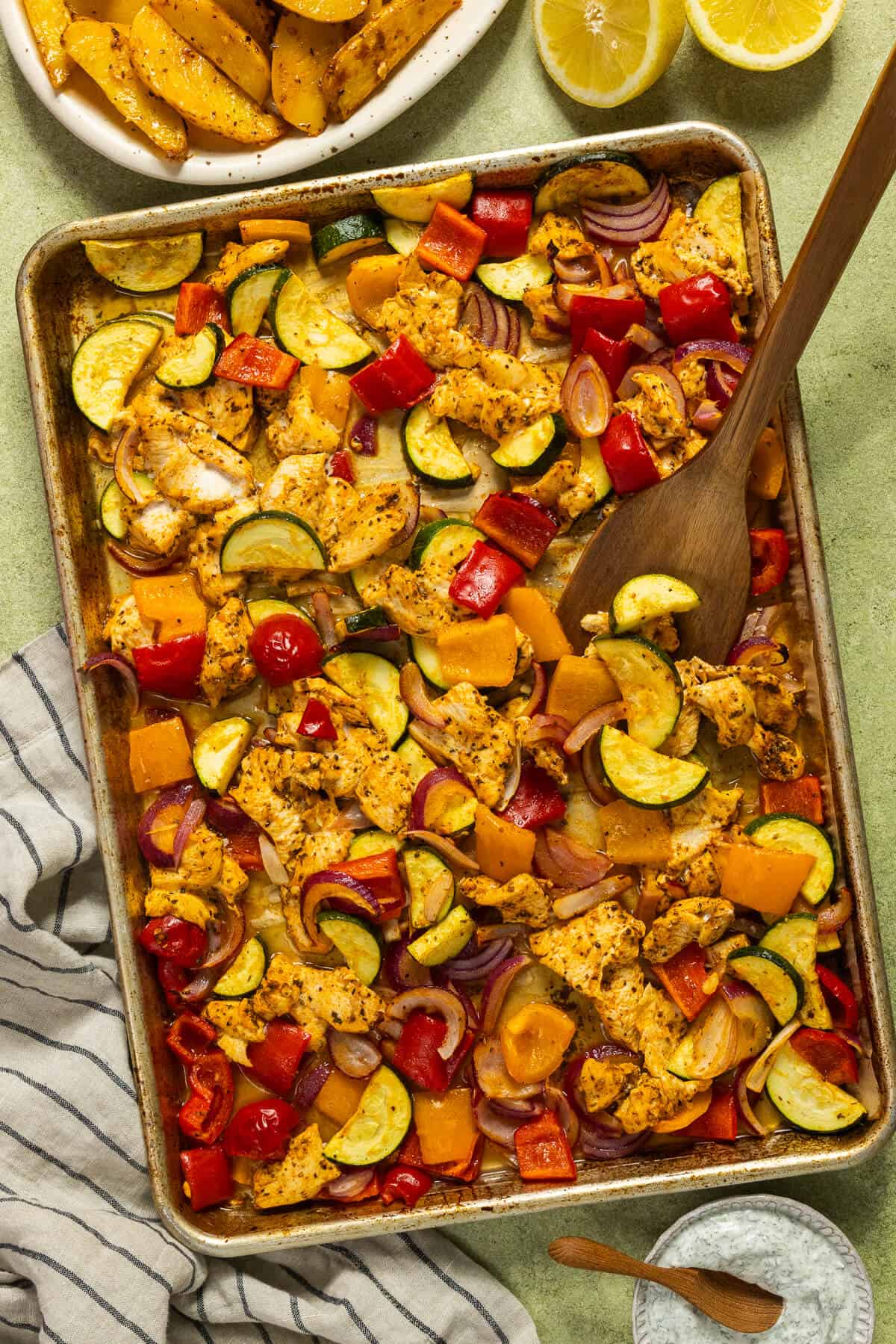 A baking sheet and wooden serving spoon with greek sheet pan chicken and vegetables.
