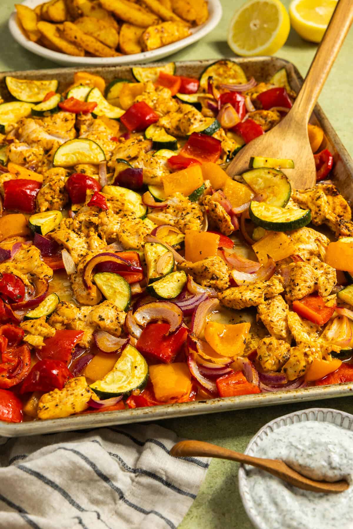 A baking sheet and wooden spatula serving the greek sheet pan chicken and vegetables.